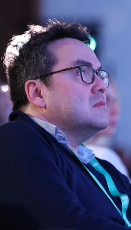 A man with dark hair and glasses wearing a blue lanyard and dark sweater listens intently, looking slightly upward.