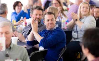 People seated at a social event clap and interact with one another. Some wear name badges and casual clothing, and tables are set with condiments and drinks.