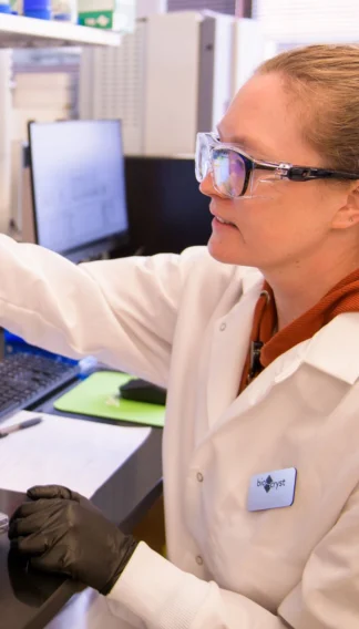 A lab technician wearing safety glasses and a lab coat works with equipment on a lab bench, focused on a task.