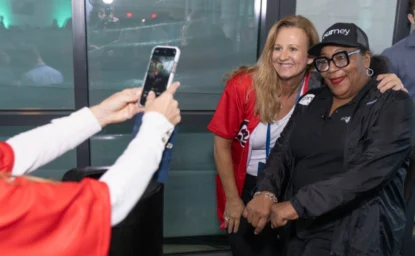 Two women posing for a photo, one in a red shirt and the other in black attire and glasses, while a third person takes the picture using a smartphone.